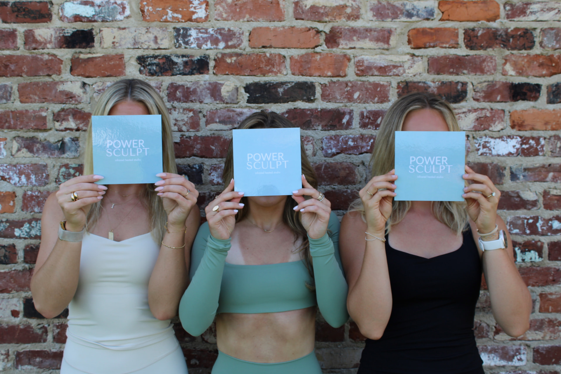 Three Power Sculpt instructors holding studio cards against a brick wall